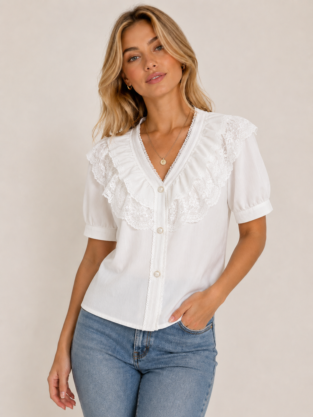 A woman wearing a white short-sleeve blouse with ruffled lace trim and button-front detailing, paired with blue jeans, against a plain beige studio background.