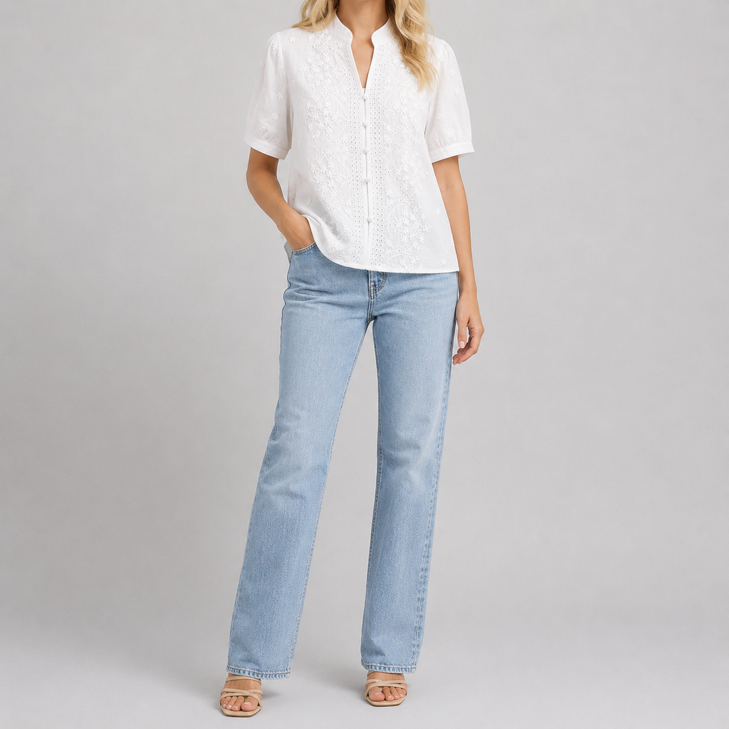 A woman modeling a white short-sleeve button-up blouse with a textured pattern and light-wash blue straight-leg jeans, standing against a neutral gray studio background.