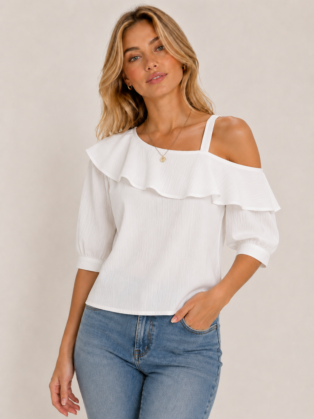 A woman wearing a white cold-shoulder top with a ruffled collar and puff sleeves, paired with blue jeans, against a plain beige background.