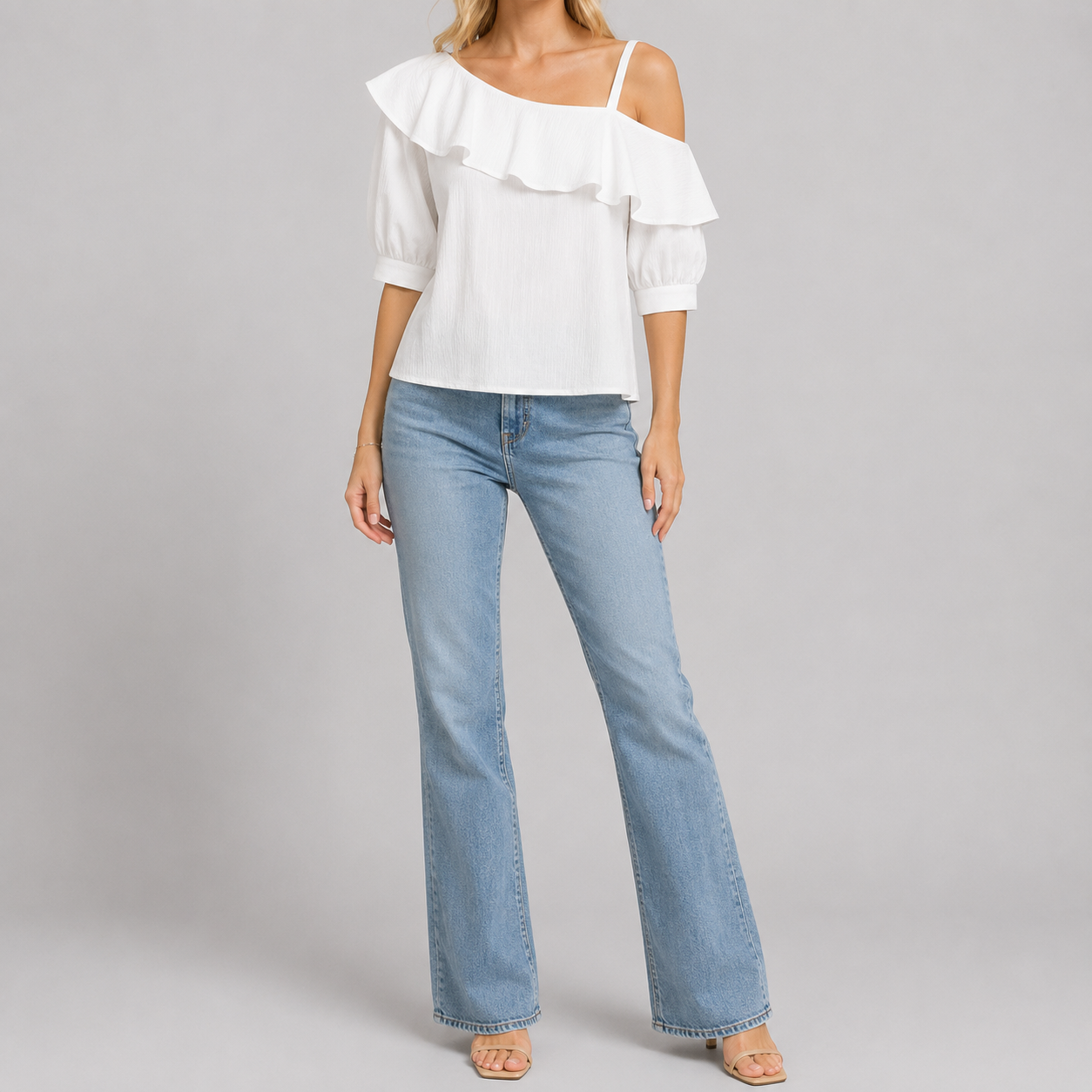 A woman modeling a white cold-shoulder top with ruffle trim and light-wash blue flare jeans against a plain light gray studio background.
