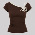 A dark brown short-sleeve top with an asymmetrical cream-colored neckline and a decorative cream bow detail on the chest, displayed on a neutral gray background.